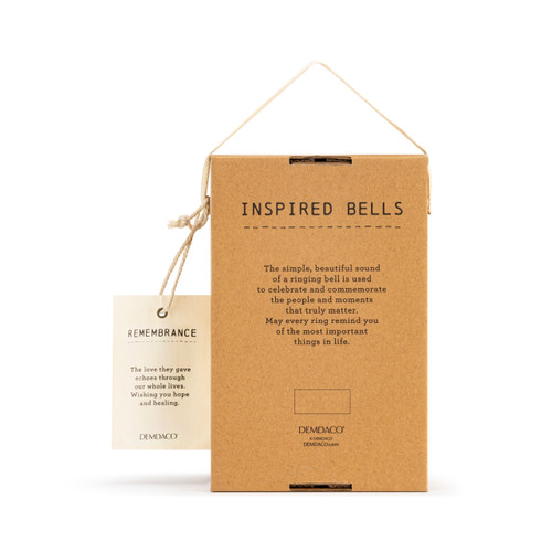 The backside of a cream Inspired Bell" with a single crack, small gray font coming from the bottom edge, a twine rope, and silver and wooden beads. Placed in a brown cardboard box with brown crinkle paper, and an ivory tag that reads "Remembrance"."