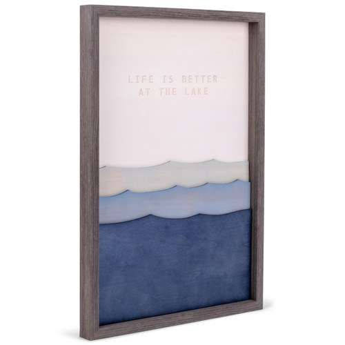 A gray frame enclosing a blue lake waves wall art piece that reads "life is better at the lake", displayed angled to the right.