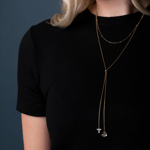 Close up of a woman wearing a black top and a long gold chain lariat necklace with a cross and heart charm at the bottom.