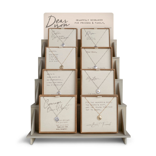 A gray wooden shelved displayer, filled with an assortment of 8 "Dear You" necklaces, each placed in a brown cardboard packaging.