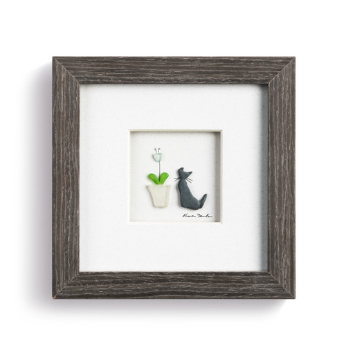 A pebble scene of a cat sitting by a flower, enclosed in a dark wooden frame.