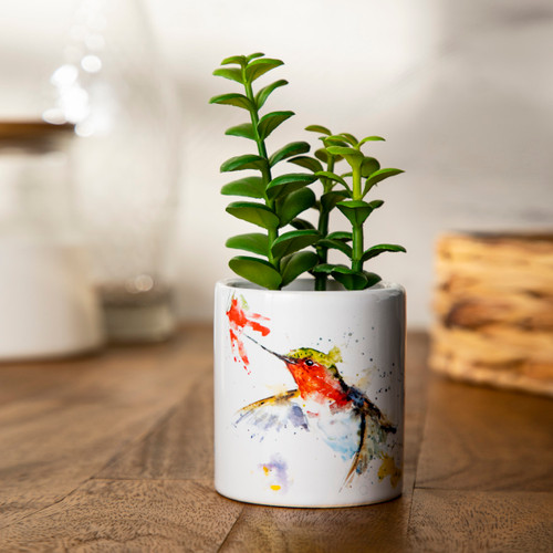 A white ceramic planter with a watercolor hummingbird and flower with a green artificial succulent inside the planter, displayed on a wood table setting.