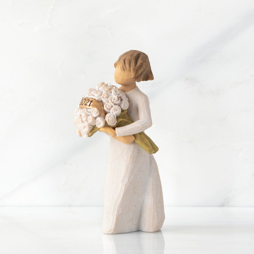 Side view, standing female figure in cream dress holding bouquet of white roses, year 2021 carved on gold-leaf plaque in bouquet