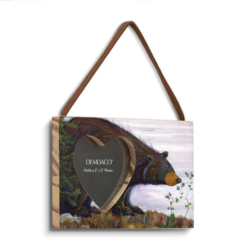 A rectangular wood hanging frame with a 2x2 inch heart shaped photo opening and has a painted image of a black bear sniffing huckleberries, displayed angled to the right.