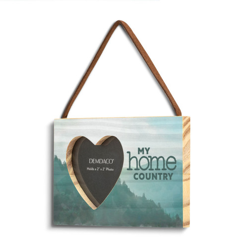 A rectangular wood hanging ornament with a 2 inch heart shaped opening for a photo next to the saying "My home country" on a green mountain background, displayed angled to the left.