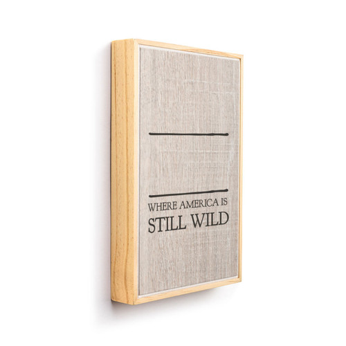 A light wood framed wall art that says "Where America is Still Wild" on a wood grain background under two black lines for personalization, displayed angled to the right.