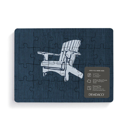 A rectangular wood 24 piece postcard puzzle with a blue Adirondack chair on a dark background with a product information tag attached.