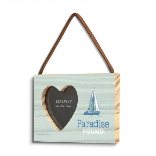 A light green rectangular wood hanging frame with a 2 inch heart shaped opening for a photo next to an image of a sailboat and the saying "Paradise Found" shown on an angle to the left.