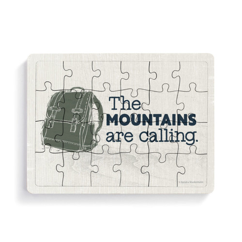 A rectangular wood 24 piece postcard puzzle with an image of a backpack and the saying "The Mountains are calling."