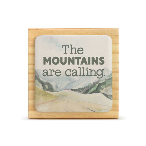 A square wood plaque that has a tile with a mountain scene and says "The Mountains are calling".
