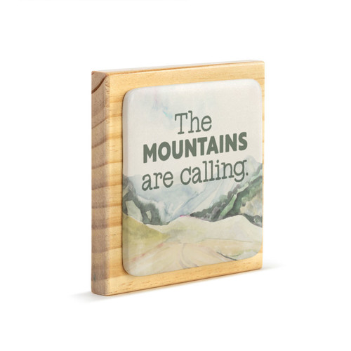 A square wood plaque that has a tile with a mountain scene and says "The Mountains are calling", displayed angled to the right.