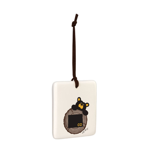 A white square hanging ornament with a black bear peeking over a tree stump with Colorado on it, displayed angled to the right.