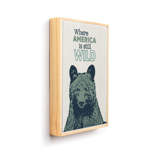 A graphic art canvas print of a bear's head that says "Where America is still Wild" above it in a light wood frame angled to the right.