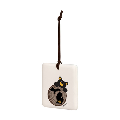 A white square hanging ornament with a black bear peeking over a tree stump with Michigan on it, displayed angled to the left.