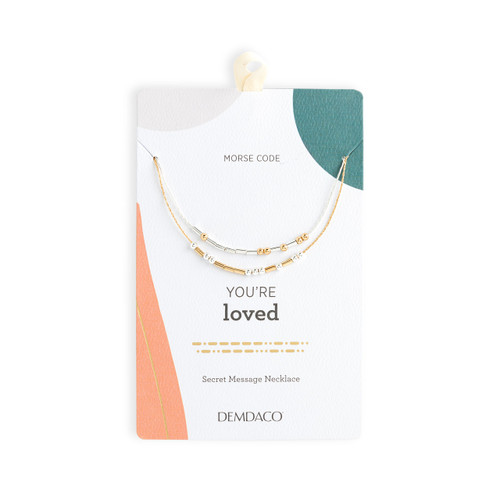Silver and gold necklace with rods and beads spelling out morse code with the theme of being loved, shown on a packaging backer card.