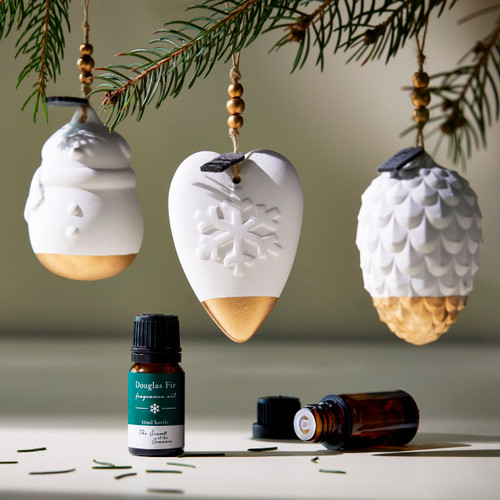 Three different white diffuser holiday ornaments hanging from a tree with two essential oil bottles on the table below.