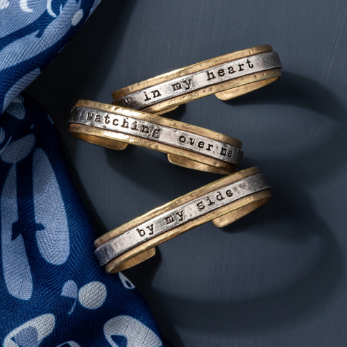 Three different mixed metal cuff bracelets. Each has a different inspirational saying and they are displayed on a dark blue background, next to a blue scarf.