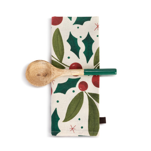 A cream kitchen towel with holly leaves and red berries next to a wood spoon with a green handle, displayed with the towel folded and the spoon laid horizontally across it.
