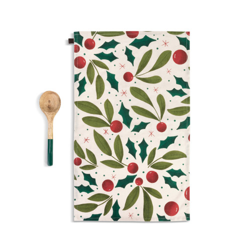 A cream kitchen towel with holly leaves and red berries next to a wood spoon with a green handle, shown with the towel laid out in full.