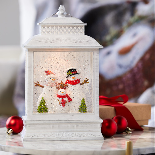 A white lit musical lantern with a family of snowmen inside, displayed on a white marble tabletop.