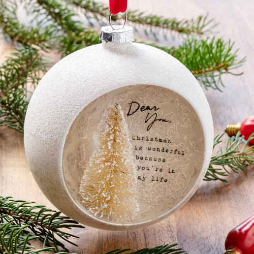 A white round cloche ornament with a white frosted tree and a "Dear You" encouraging message inside, displayed over a wood surface with evergreen branches and red Christmas lights.