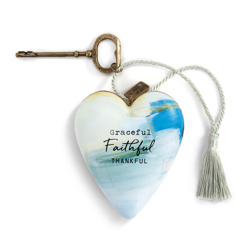 A heart shaped sculpture with a gold key and silver tassel featuring artwork created by ArtLifting artist Charlie French. The image is light and darker blue paint stripes and says "Graceful Faithful Thankful".