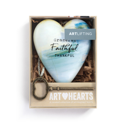 A heart shaped sculpture with a gold key and silver tassel featuring artwork created by ArtLifting artist Charlie French. The image is light and darker blue paint stripes and says "Graceful Faithful Thankful", displayed in a packaging box.