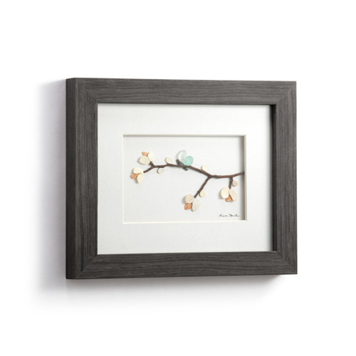 A gray wood framed image of a small tree branch with a butterfly and flowers made of pebbles, displayed angled to the right.