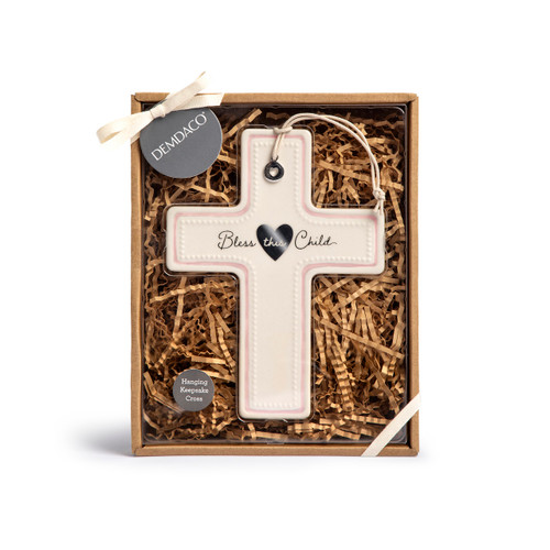 A cream ceramic hanging cross with a pink accent and says Bless this Child with a silver token at the top, displayed in a packaging box.