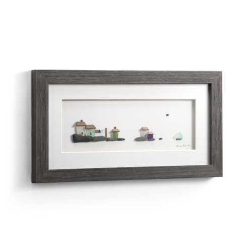 A gray wood framed image of a little village by the sea made of pebbles, displayed angled to the right.