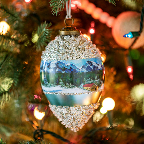 An onion shaped hanging ornament with an image of a winter Swiss Chalet around the middle and silver glitter around the top and bottom inspired by artwork from ArtLifting artist Lucas Farlow, displayed hanging in a lit Christmas tree.