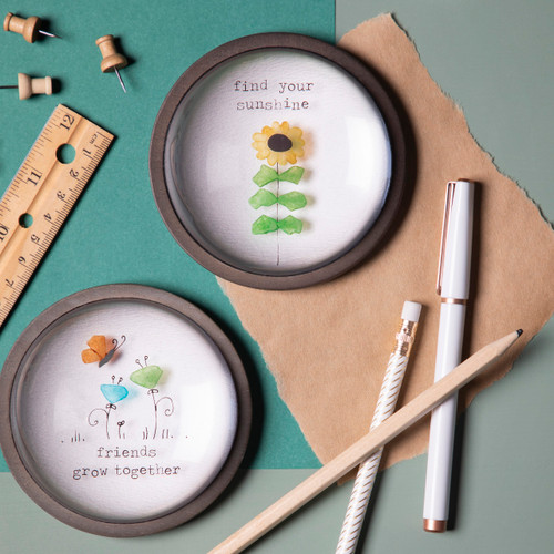 Two different round paperweights with images made of colored stone. One is a sunflower and says "find your sunshine" the other is flowers and says "friends grow together".