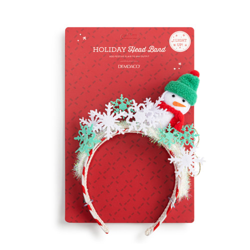 A fun red and white holiday snowflake headband with a small snowman in a green hat on top, displayed on a packaging backer card.