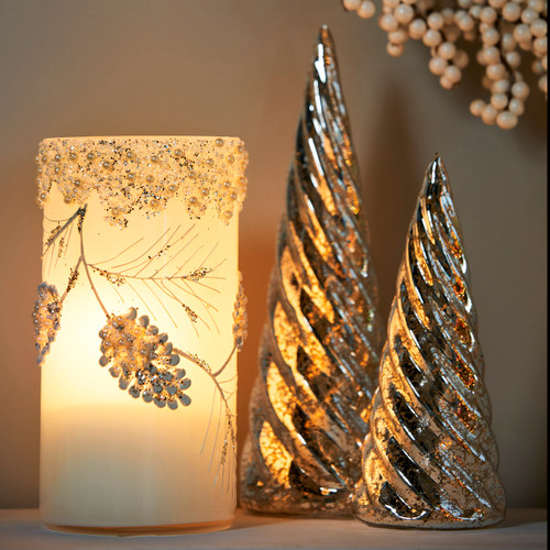 A lit cylinder decorated on the outside with glitter leaves and branches, displayed next to two silver sculpted trees.