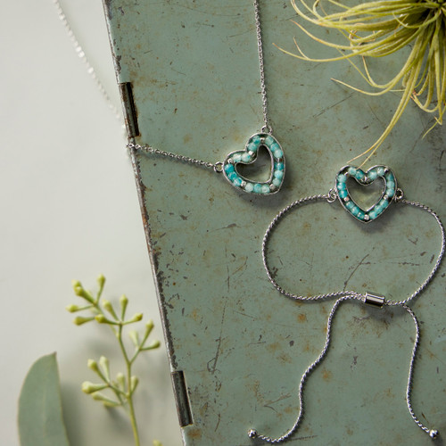 A silver chain bracelet and necklace each with a heart shaped charm filled with aqua beads, displayed on a green metal box with assorted greenery.