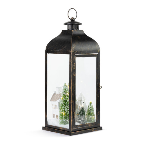 A lit black metal lantern with a winter village scene inside with snow and evergreens, displayed angled to the right.