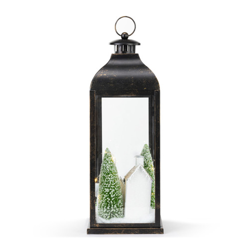 Back view of a lit black metal lantern with a winter village scene inside with snow and evergreens.
