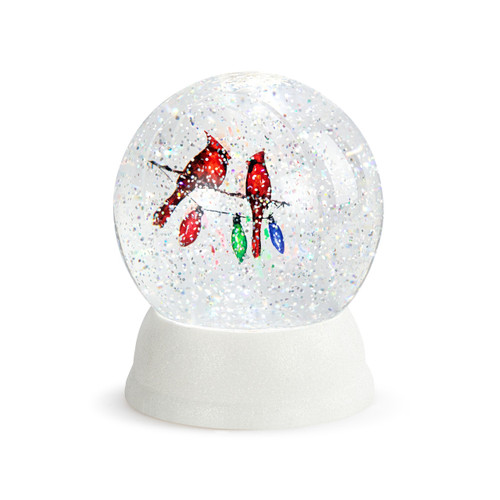 A white round glass snow globe with the image of two cardinals on a string of holiday lights, displayed angled to the left.