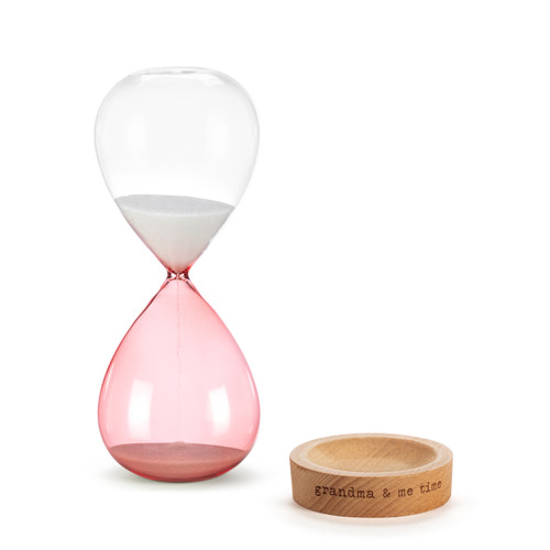 A glass sand timer with pink glass on the bottom half sitting in a wood base that reads "grandma & me time", displayed with the glass timer off the base.