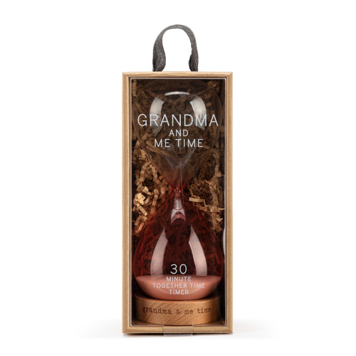 A glass sand timer with pink glass on the bottom half sitting in a wood base that reads "grandma & me time", displayed in a packaging box.