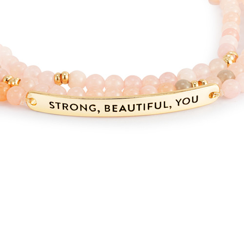 Detail view of the gold metal bar on a multi-strand beaded bracelet in a mix of pink with a gold metal bar that says "Strong, Beautiful, You".