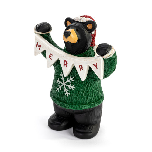 A sculpted figurine of a black bear wearing a green and white snowflake sweater, a Santa hat and holding a white garland that says "Merry", displayed angled to the left.