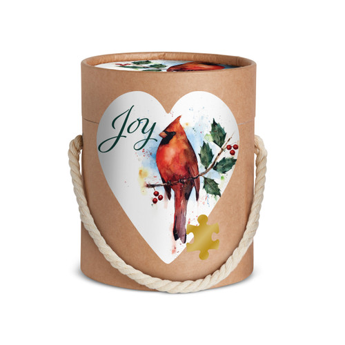 The cardboard tube packaging with the artwork for a heart shaped 100 piece puzzle of a red cardinal perched on a holly branch that says "Joy".