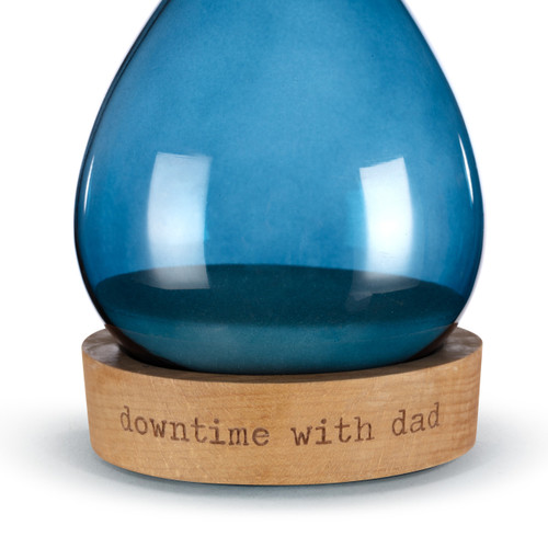 Detail view of the wood base on a glass sand timer with blue glass on the bottom half sitting in a wood base that reads "downtime with dad".