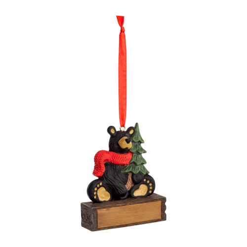 A hanging ornament with a sitting black bear in a red scarf and holding a pine tree on a rectangular base that can be personalized, displayed angled to the right.