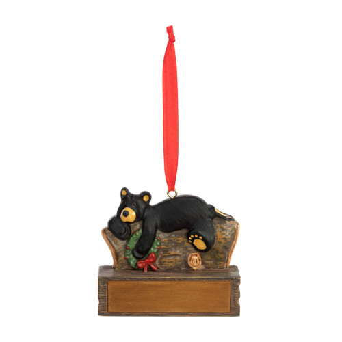 An ornament of a black bear laying on a log and holding a wreath, hanging from a red ribbon. There is a spot in front for customization.