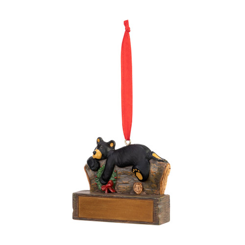 An ornament of a black bear laying on a log and holding a wreath, hanging from a red ribbon. There is a spot in front for customization, displayed angled to the left.