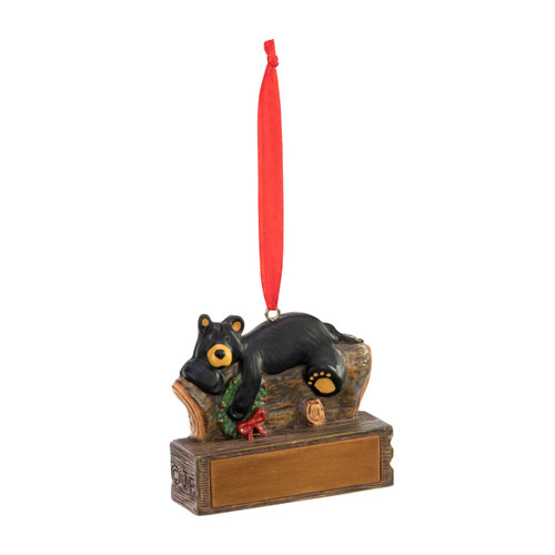 An ornament of a black bear laying on a log and holding a wreath, hanging from a red ribbon. There is a spot in front for customization, displayed angled to the right.