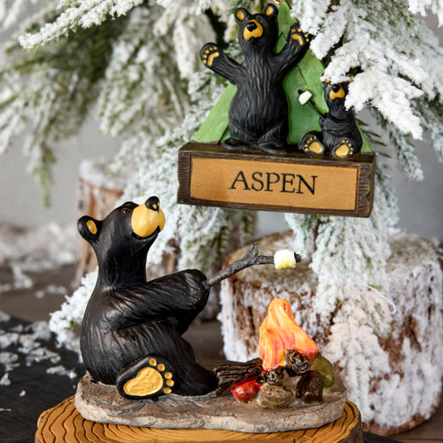 A figurine of a black bear toasting marshmallows over a campfire. There is also a hanging ornament of two black bears. Each has a place to personalize the figure. The figure and ornament are displayed on a table surface with a decorative frosted tree.