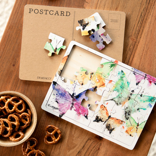 Overhead view of a partially completed postcard puzzle of colorful butterflies, laid on a wood table next to some snacks.